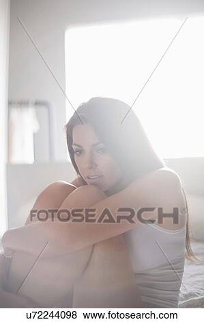 Stock Photo - Young woman sitting on bed, hugging knees. Fotosearch