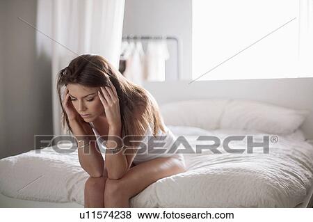 Young woman sitting on edge of bed, holding head View Large Photo Image Stock Photo - Young woman sitting on edge of bed, holding head. Fotosearch