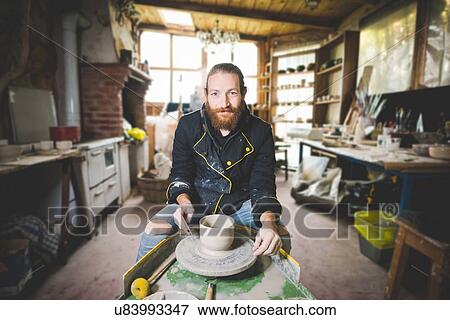 Stock Photo - Bearded mid adult man in workshop sitting at pottery wheel making clay pot, looking at camera smiling. Fotosearch
