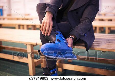 Stock Image - Chest down of mid adult man sitting on bench putting on ice skates. Fotosearch