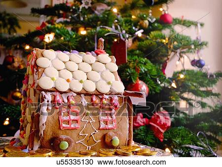 Stock Image - Confectionery covered gingerbread house in front of christmas tree. Fotosearch