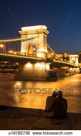 Stock Photograph - Couple on the Danube bank, Chain Bridge in background, at night, Hungary, Budapest. Fotosearch