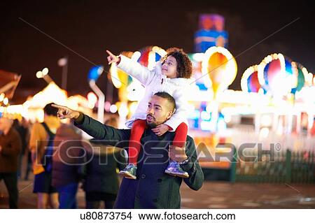 Picture - Father carrying daughter on shoulders at funfair, father and daughter pointing. Fotosearch