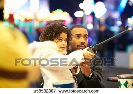 Stock Photo - Father helping daughter with rifle at shooting gallery at funfair. Fotosearch