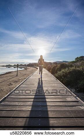 Stock Photograph - Female runner and her shadow running along beach boardwalk, Villasimius, Sardinia, Italy. Fotosearch