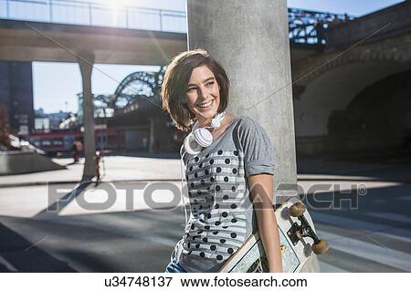 Stock Photo - Female skateboarder with skateboard leaning against urban pillar. Fotosearch
