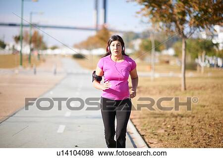 Stock Photo - Front view of mature woman wearing headphones and armband jogging on pathway. Fotosearch