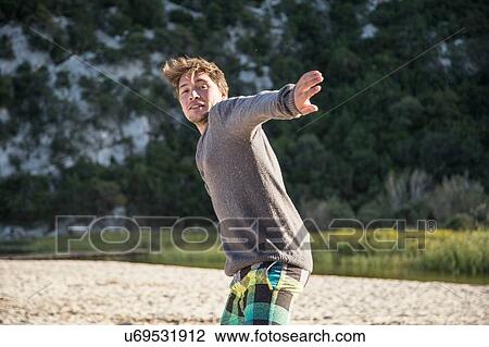Stock Image - Front view of young man on beach poised in throwing action, looking at camera smiling, Costa Smeralda, Sardinia, Italy. Fotosearch