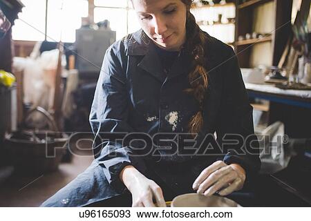 Stock Image - Front view of young woman sitting at pottery wheel looking down making clay pot. Fotosearch