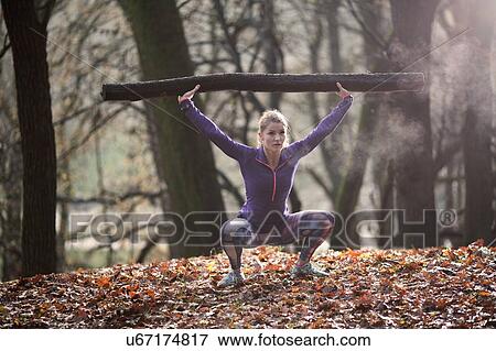 Stock Photo - Front view of young woman squatting in forest arms raised holding tree branch. Fotosearch