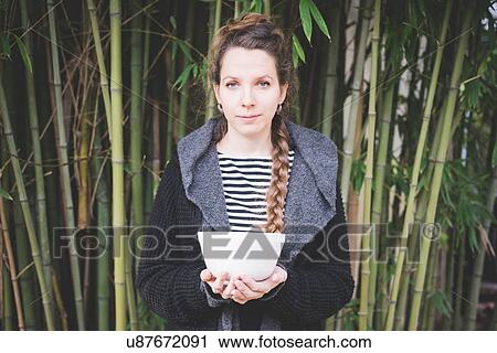 Stock Image - Front view of young woman standing in front of bamboo grove holding ceramic dish looking at camera. Fotosearch