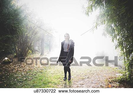 Stock Image - Full length front view of young woman standing in misty garden looking at camera smiling. Fotosearch