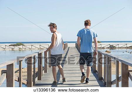 Stock Photograph - Full length rear view of young men walking on wooden pier, looking over shoulder, Sardinia, Italy. Fotosearch