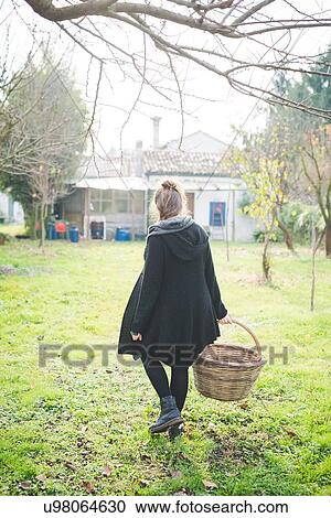 Stock Image - Full length rear view of young woman in garden carrying wickerwork basket. Fotosearch