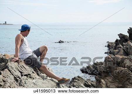 Full length side view of young man sitting on rocks looking away at ocean, Stintino, Sardinia, Italy View Large Photo Image Stock Image - Full length side view of young man sitting on rocks looking away at ocean, Stintino, Sardinia, Italy. Fotosearch