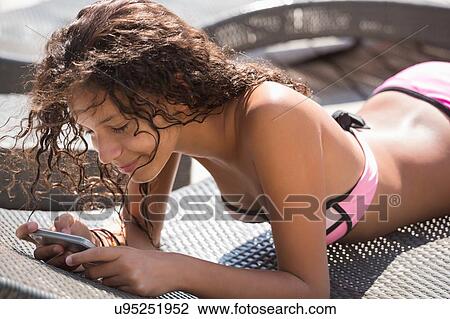 Stock Image - Girl lying on sunbed reading smartphone texts. Fotosearch