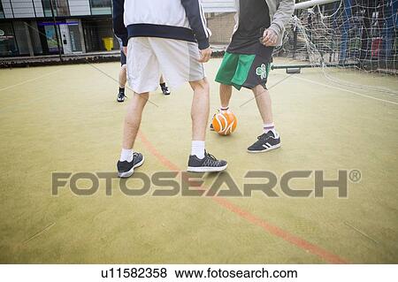 Stock Photo - Group of adults playing football on urban football pitch, low section. Fotosearch