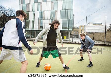 Stock Photography - Group of adults playing football on urban football pitch. Fotosearch