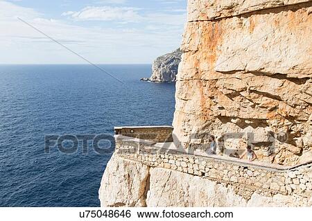 Stock Photograph - High angle view of young men by ocean on balcony carved into cliff, Grotta di nettuno (Neptune's Grotto), Capo Caccia, Sardinia, Italia. Fotosearch