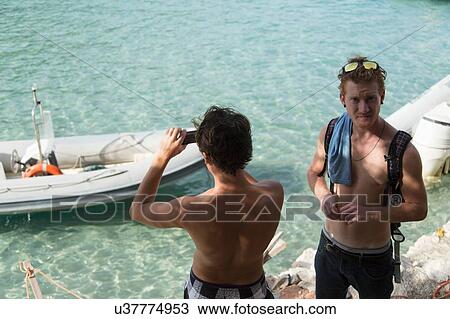 Stock Image - High angle view of young men using smartphone to photograph boat on clear water, Cala Luna, Sardinia, Italy. Fotosearch
