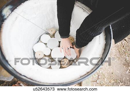 Stock Image - High angle view of young woman arms reaching into barrel kiln for clay pot. Fotosearch