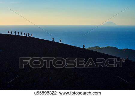 Stock Image - Large group of silhouetted tourists walking rim at Gran Cratere, Vulcano Island, Aeolian Islands, Sicily, Italy. Fotosearch