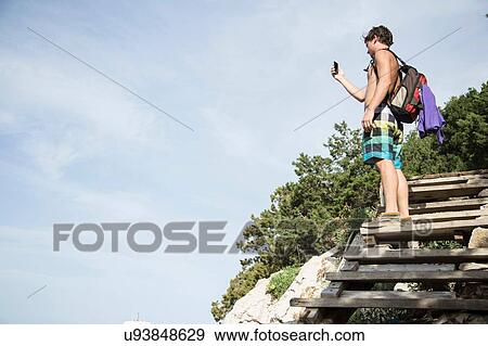 Low angle side view of young man standing on wooden steps using smartphone to take photograph, Cala Luna, Sardinia, Italy View Large Photo Image Stock Photo - Low angle side view of young man standing on wooden steps using smartphone to take photograph, Cala Luna, Sardinia, Italy. Fotosearch