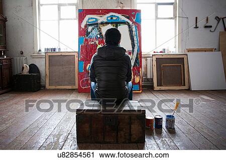 Stock Image - Male artist sitting on crate, looking at artwork, rear view. Fotosearch