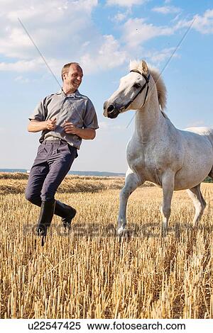 Stock Photography - Male trainer running in front of white horse in field. Fotosearch
