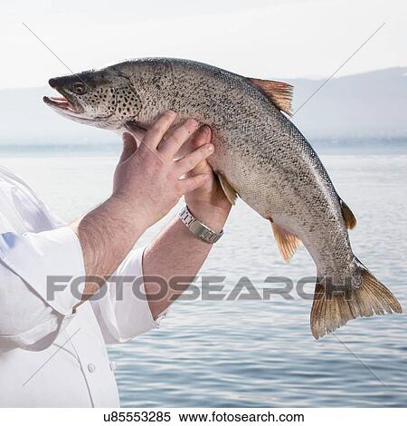 Stock Photography - Man holding up fish, lake in background. Fotosearch