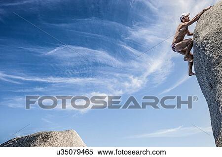 Stock Photography - Man wearing swimming trunks climbing rock, Villasimius, Sardinia, Italy. Fotosearch