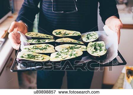 Stock Photo - Mans hands holding aubergine halves on baking tray in kitchen. Fotosearch