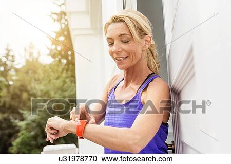 Stock Image - Mature female runner checking smartwatch at front door. Fotosearch