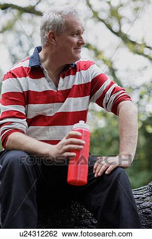Stock Image - Mature male sitting on forest tree trunk taking a water break. Fotosearch