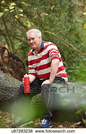 Stock Photo - Mature male sitting on forest tree trunk drinking from water bottle. Fotosearch