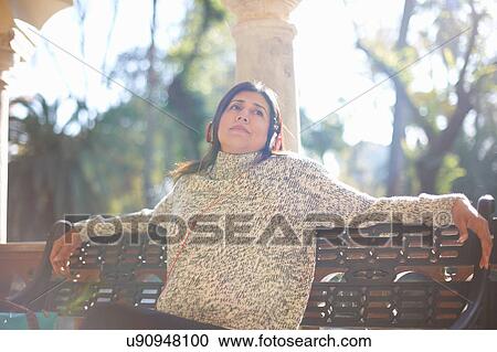 Stock Image - Mature woman sitting bench wearing headphone looking away, Seville, Spain. Fotosearch
