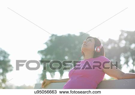 Stock Image - Mature woman sitting on park bench wearing headphones looking up. Fotosearch