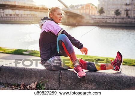 Stock Photo - Mid adult female runner doing warm up exercise on riverside wall. Fotosearch