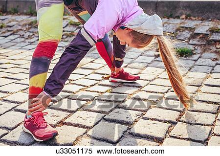 Stock Photography - Mid adult female runner doing warm up exercise on cobblestones. Fotosearch