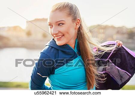 Stock Image - Mid adult female runner putting on tracksuit top at city riverside. Fotosearch