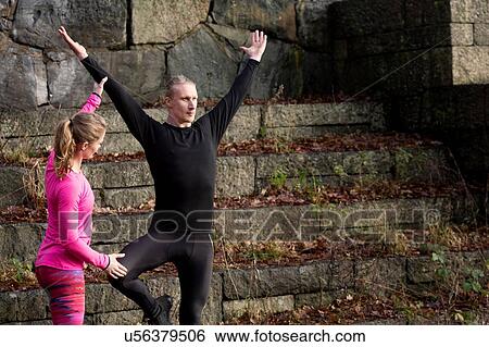 Stock Photograph - Mid adult man on stone steps arms raised standing on one leg, young woman helping. Fotosearch