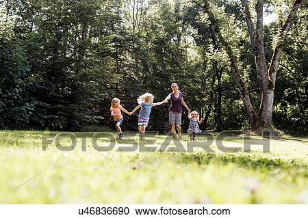 Stock Image - Mid adult woman and three young daughters holding hands and running in park. Fotosearch