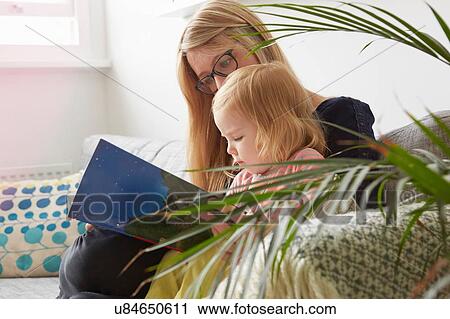 Stock Image - Mid adult woman and toddler daughter reading on living room sofa. Fotosearch