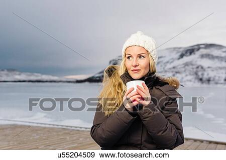 Stock Photo - Mid adult woman on pier by frozen lake holding mug looking away smiling. Fotosearch