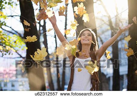 Stock Photo - Mid adult woman throwing autumn leaves in sunlit park. Fotosearch