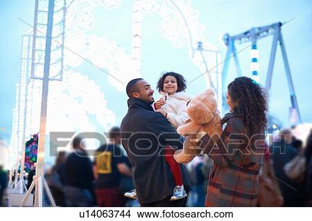 Mother and father in amusement park carrying smiling girl and teddy bear View Large Photo Image Picture - Mother and father in amusement park carrying smiling girl and teddy bear. Fotosearch