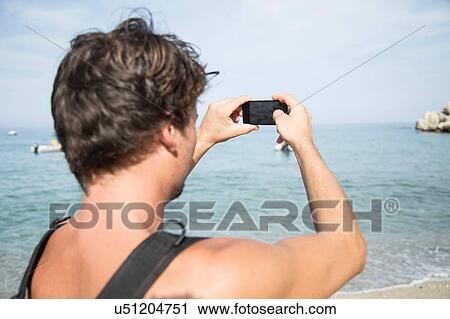 Stock Image - Over shoulder rear view of young man using smartphone to photograph horizon over water, Cala Luna, Sardinia, Italy. Fotosearch