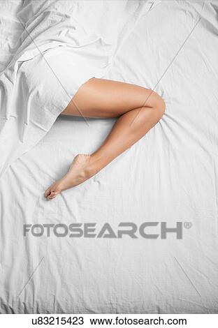 Stock Image - Overhead view of white sheets and female bare leg on bed. Fotosearch