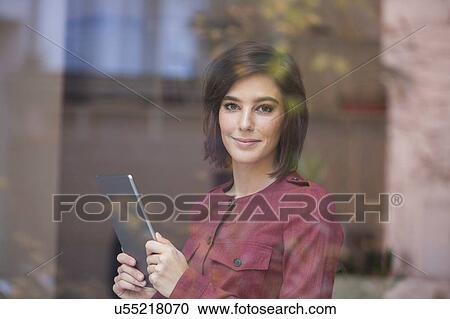 Stock Image - Portrait of businesswoman holding digital tablet looking through window. Fotosearch