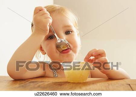 Stock Image - Portrait of female toddler eating dessert at table. Fotosearch